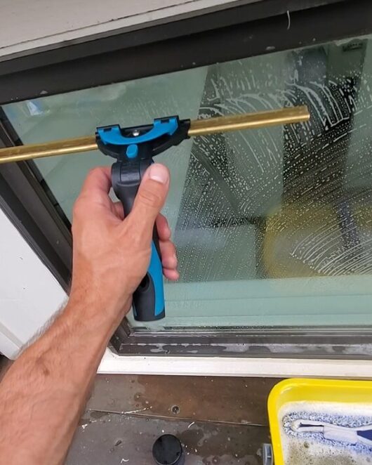 Man's arm squeegeeing a window with a yellow bucket filled with water on the ground